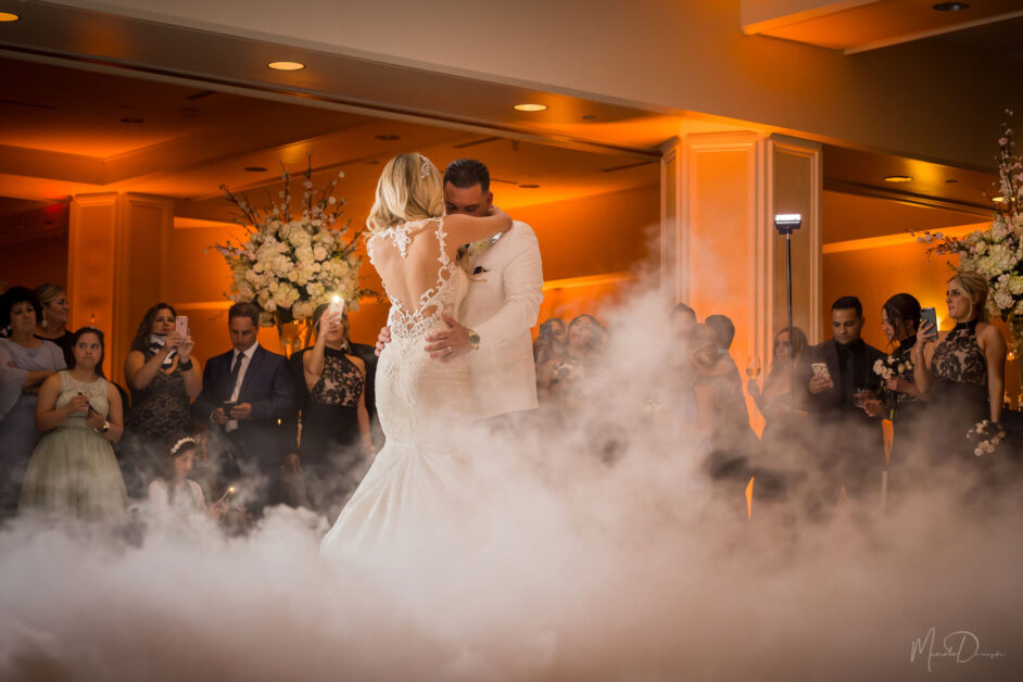 Una novia y un novio comparten un baile rodeados de niebla en una pista de baile nupcial, su audiencia cautivada por el momento, una cálida iluminación ambiental realza la atmósfera romántica. By www.powerparties.com