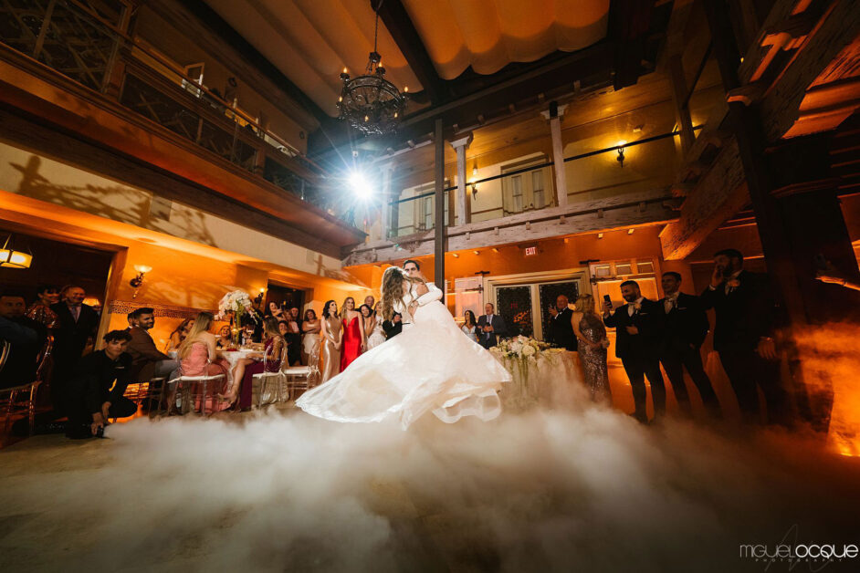 Una novia y un novio comparten su primer baile en una pista de baile cubierta de humo, rodeados de invitados en un lugar elegantemente iluminado con un brillo ambiental cálido en su boda en Miami. By www.powerparties.com
