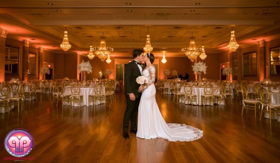 Una pareja de recién casados comparte un beso en el centro de un salón de baile elegantemente iluminado, rodeado de mesas redondas con centros de mesa florales blancos y candelabros brillantes en el techo, celebrando su boda en Miami. By www.powerparties.com