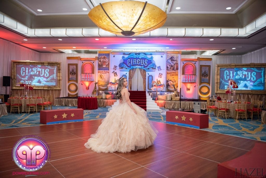 A person in a voluminous ball gown walking through a Miami Quince-themed event hall decorated with large posters, red stars, and a central chandelier. By www.powerparties.com
