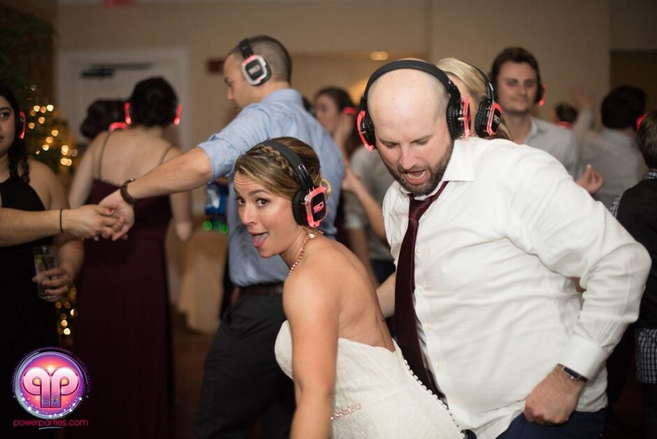 Una novia y un hombre con camisa y corbata bailan con entusiasmo en una boda disco silenciosa en Miami, ambos con audífonos rojos. Al fondo se ven otros invitados, también con auriculares. By www.powerparties.com