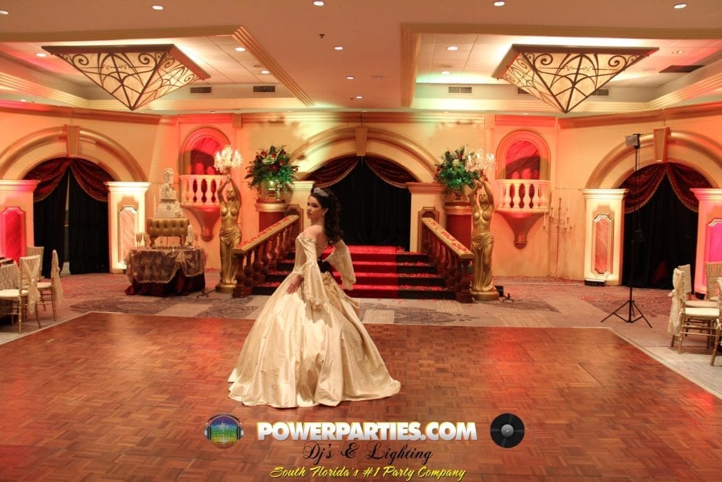 Una novia con un voluminoso vestido blanco está sola en un gran salón de baile profusamente decorado con una escalera, columnas doradas y una cálida iluminación ambiental, lista para la boda hora loca en Miami. By www.powerparties.com