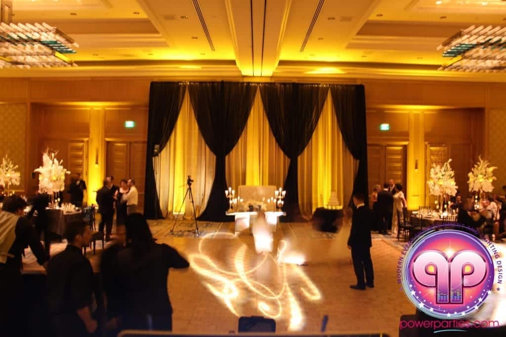 Elegante salón para eventos de bodas con invitados, mesas elaboradas, iluminación espectacular y grandes decoraciones florales con un DJ hora loca, con el logotipo de "powerparties.com" superpuesto en el By www.powerparties.com