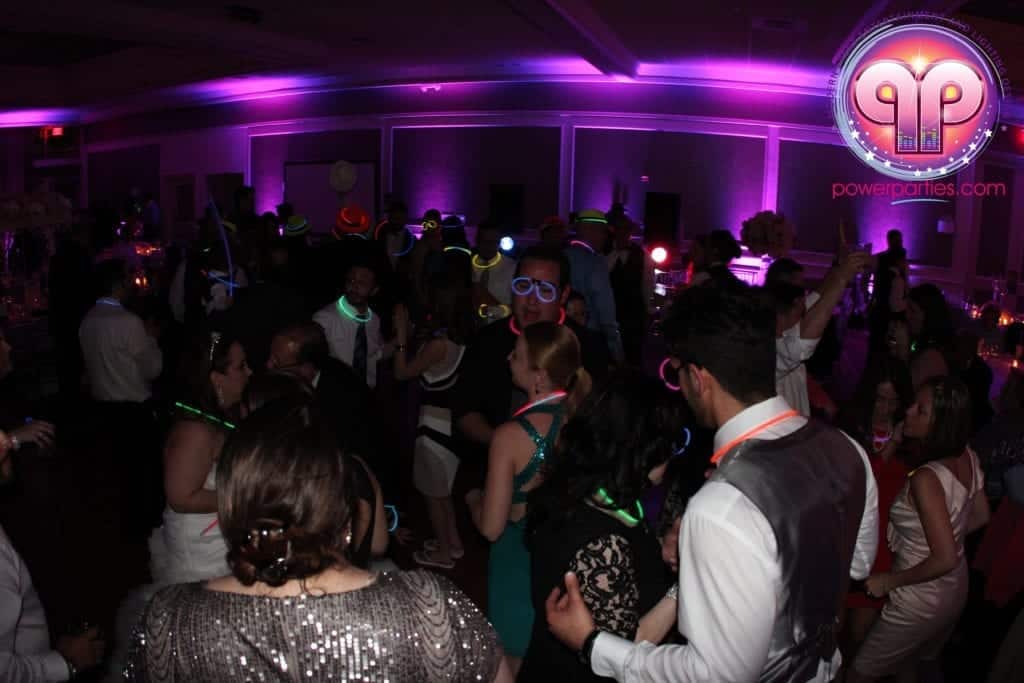 Una animada pista de baile en una fiesta de quince en Miami con invitados usando accesorios brillantes y luces de colores iluminando la sala durante la hora loca. Se muestra un logotipo para "powerparties.com" By www.powerparties.com