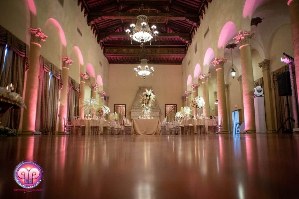 A beautifully decorated Biltmore Hotel wedding venue with ornate chandeliers, high ceiling, columns, and an elaborately set table at the center, surrounded by floral arrangements. By www.powerparties.com