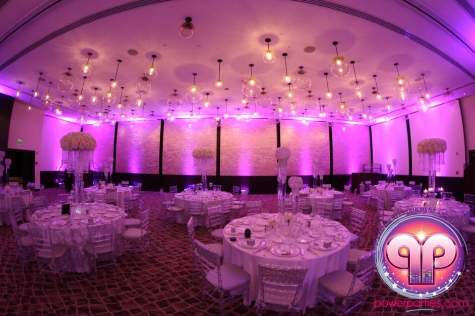 Un elegante salón de banquetes iluminado con iluminación rosa para una boda, con mesas redondas preparadas para cenar, rematadas con grandes centros de mesa florales y rodeadas de sillas blancas. Del techo cuelgan luces decorativas redondas. By www.powerparties.com