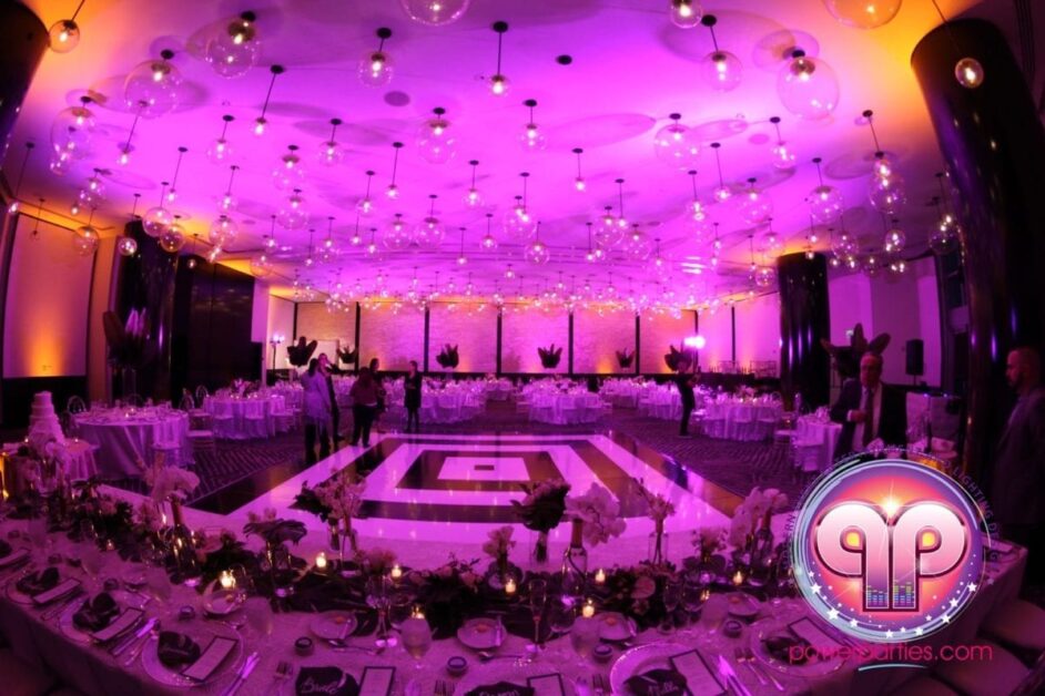 Un elegante salón de banquetes de bodas iluminado con luces violetas y azules, que cuenta con una pista de baile en el centro rodeada de mesas redondas preparadas para cenar y candelabros de cristal ornamentados en el techo. By www.powerparties.com