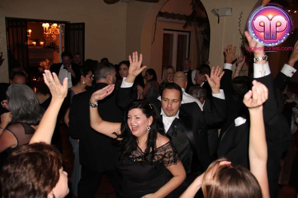 Una animada fiesta de boda protagonizada por la "hora loca" con los adultos bailando y levantando las manos alegremente en un salón con decoración tradicional. En primer plano, una mujer sonríe ampliamente mientras baila. By www.powerparties.com