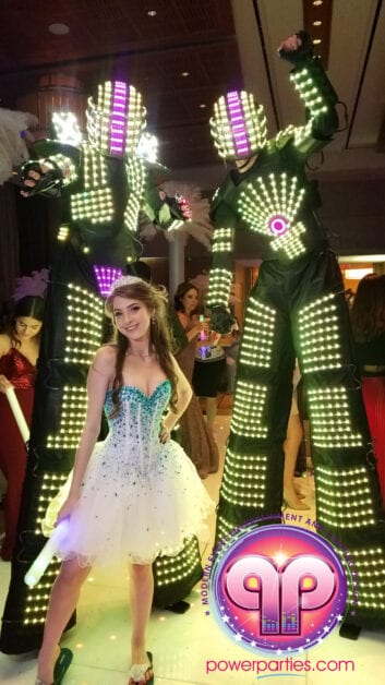 A woman in a sparkling white dress and tiara stands between two figures covered in illuminated green LED lights at a festive hora loca event, with event logos visible. By www.powerparties.com
