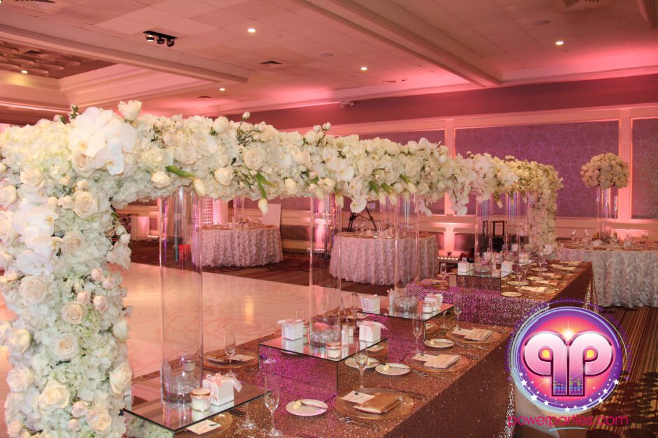 Un elegante salón de recepción de bodas en Miami adornado con un gran arco floral de flores blancas. El lugar cuenta con iluminación rosa, mesas con espejos y vajilla fina y una pista de baile personalizada con un logotipo. By www.powerparties.com