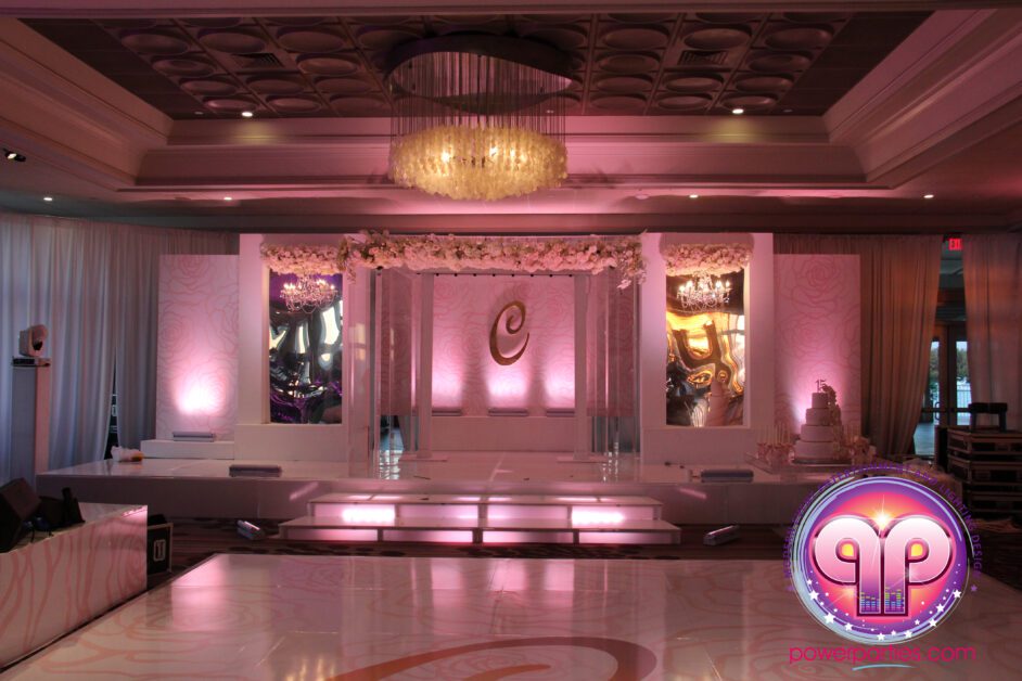 Elegant Rusty Pelican Quince setup with a lit stage featuring intricate floral patterns, an elaborate monogram, and ambient pink lighting under a luxurious chandelier. By www.powerparties.com