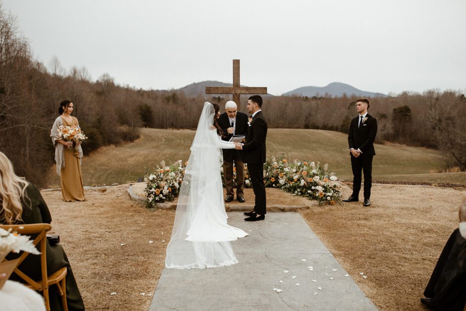 A destination wedding ceremony outdoors in Georgia with a couple exchanging vows at an altar under a wooden cross, framed by mountains and a cloudy sky. Guests and a bridal party observe the moment. By www.powerparties.com