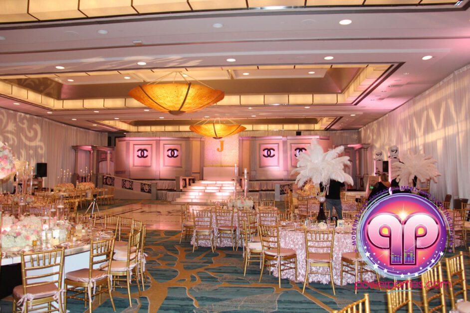 Elegant event hall decorated for a Chanel Quince with round tables, gold chairs, pink lighting, large white feather centerpieces, and ornate hanging lamps, all arranged on a blue patterned carpet By www.powerparties.com
