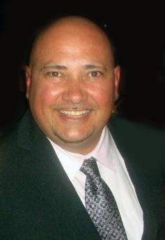 A portrait of a smiling bald man wearing a dark blazer, white shirt, and patterned tie. The background is dimly lit. By www.powerparties.com