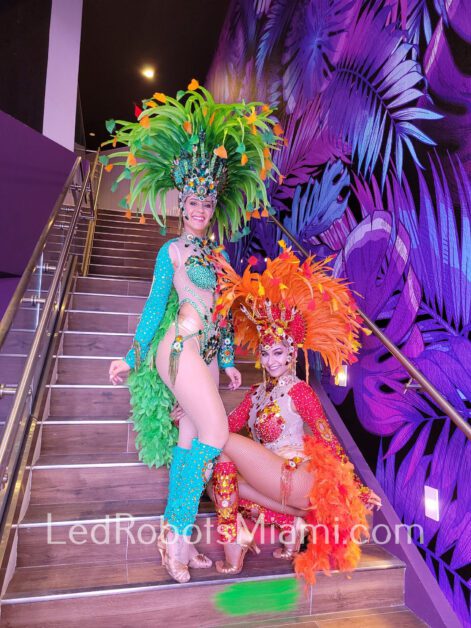 Two hora loca dancers in vibrant carnival costumes featuring feathers and sequins pose on a staircase, with large purple feather decorations in the background. By www.powerparties.com