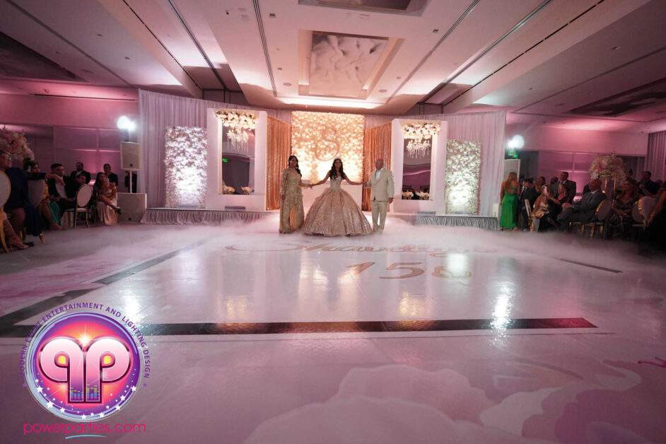 A quinceañera shares her first dance on a reflective dance floor with her initials projected onto it, surrounded by guests and elegant decor in a lavishly lit ballroom. By www.powerparties.com