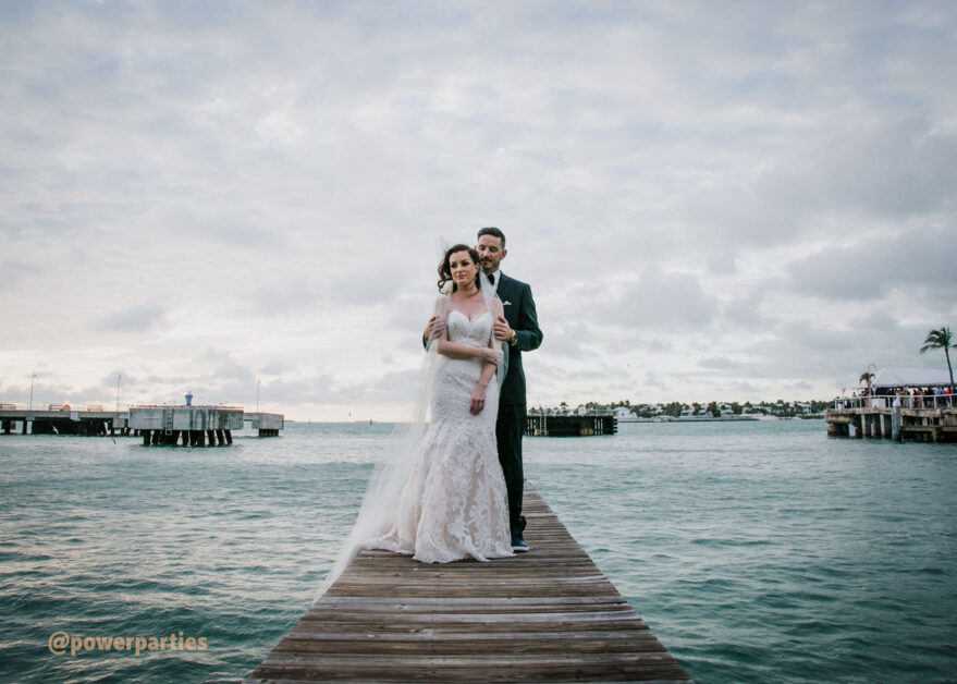 A newlywed couple embracing on a wooden pier in Key West over the ocean at dusk, with cloudy skies and calm waters in the background. The bride wears a white lace dress and the groom is in By www.powerparties.com