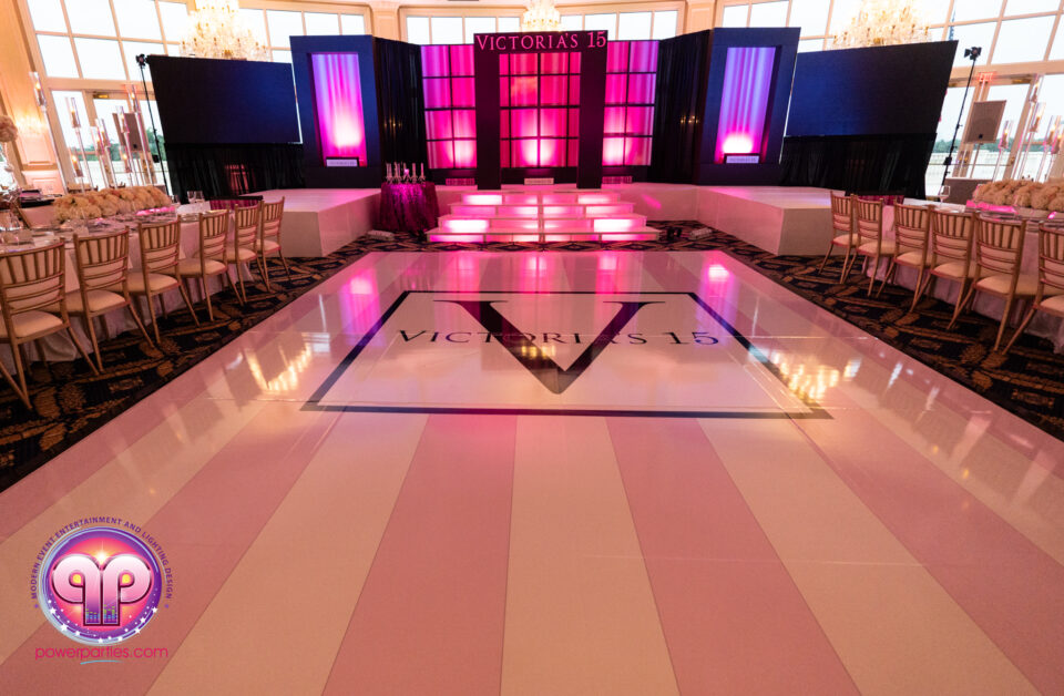 Elegant ballroom decorated for a celebration, featuring a glossy white dance floor with "victoria's 15" printed on it, surrounded by round tables set for dining, and a vibrant pink and purple light display on stage. By www.powerparties.com