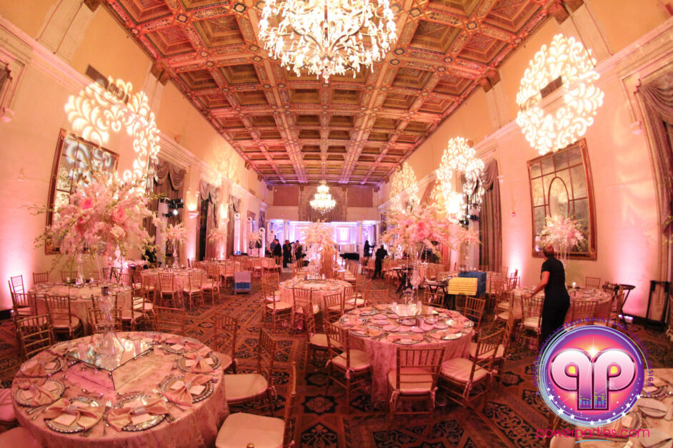 Elegant event hall at the Biltmore Hotel adorned with ornate chandeliers, pink lighting, and large floral centerpieces on round tables. The room features intricate wall and ceiling details, providing By www.powerparties.com