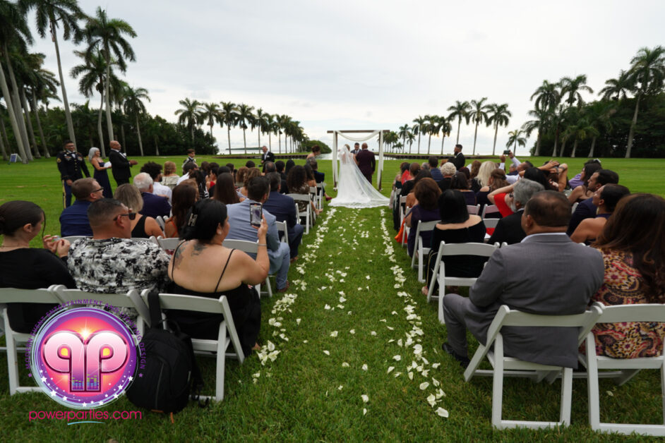 Outdoor wedding ceremony at Deering Estate with guests seated on white chairs facing a couple under a white canopy, lined by a petal-covered pathway, set against a backdrop of towering palm trees and lush green By www.powerparties.com