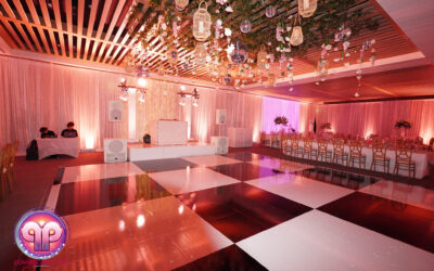Elegant wedding event space with a glossy checkered dance floor, pink ambient lighting, chairs lined up in rows, and ceiling draped with hanging floral decorations and lanterns. By www.powerparties.com