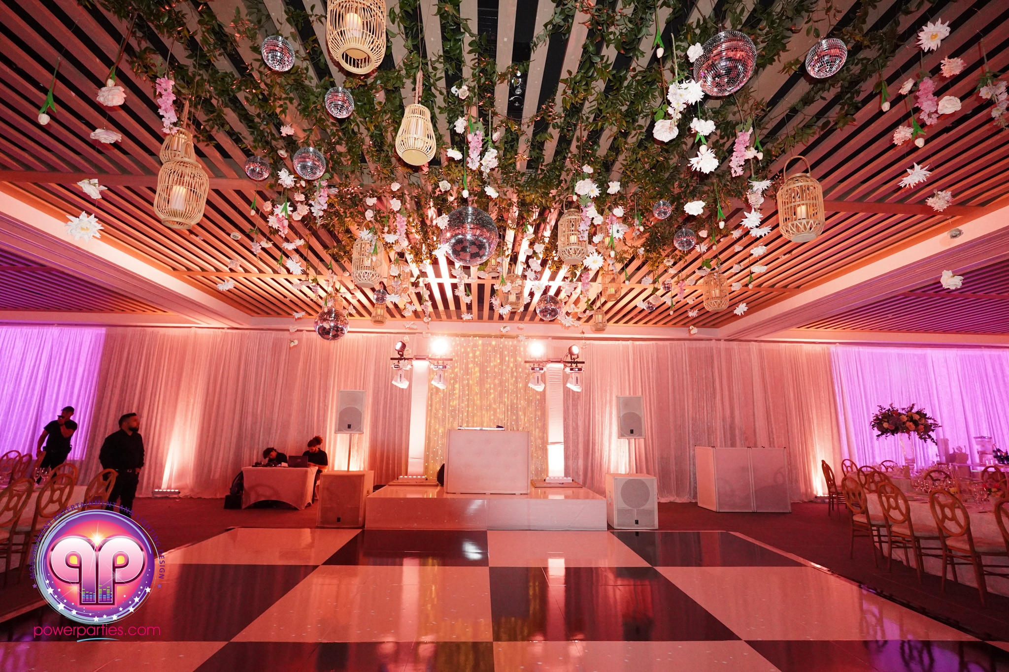 Elegant wedding reception at The Edition Miami Beach with vibrant blush lighting. the ceiling is adorned with wooden slats and hanging floral arrangements, complemented by suspended ornate lanterns. tables set for guests with a lit central stage. By www.powerparties.com