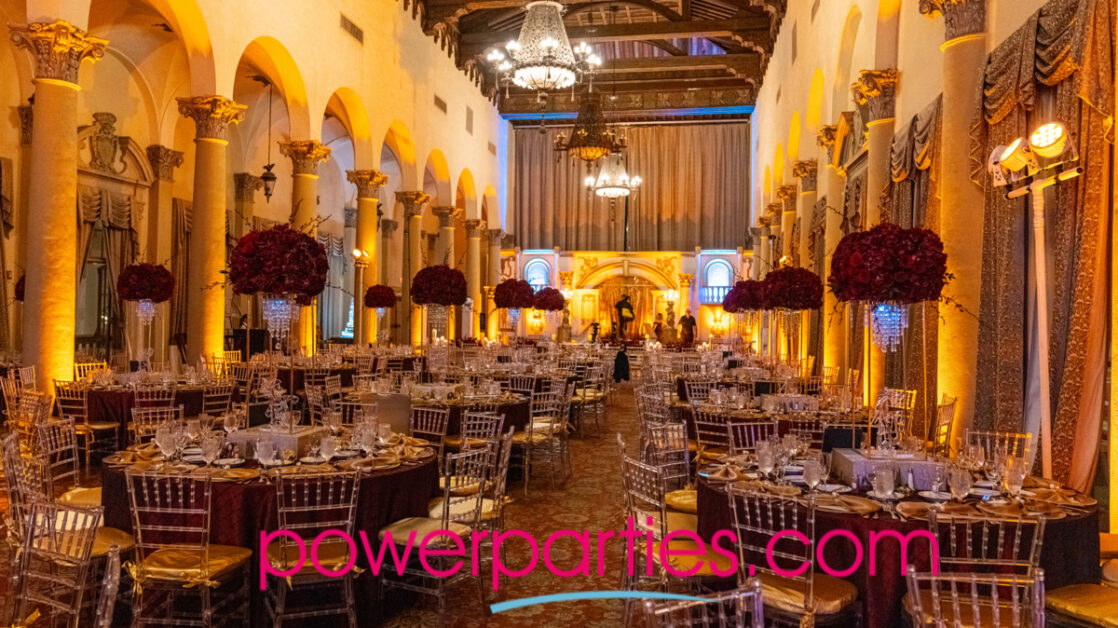 Elegant Biltmore Hotel Cora Gables ballroom with tables set for a quince, featuring crystal glassware and floral centerpieces under warm lighting, with a sophisticated archway design in the background. By www.powerparties.com