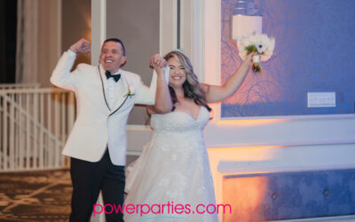 Produced by Power Parties DJs A joyful bride and groom triumphantly enter a ballroom, the groom in a white tuxedo raises his fists in excitement, and the bride in a lace gown lifts her bouquet, both smiling. a banner with "powerparties.com" is visible.