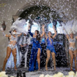 A vibrant scene with four performers in elaborate, feathered costumes, dancing amidst a shower of confetti. Two women on the left wear white costumes, another two on the right in blue. A festive atmosphere with decorations and an archway in the background. By www.powerparties.com